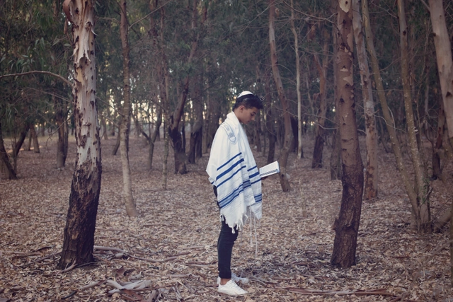A boy in a tallit and kippah stands in a forest, reading from a book.