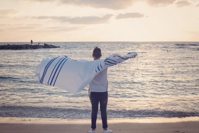 A boy stands on a beach with a tallit spread out, facing the sea at sunset.